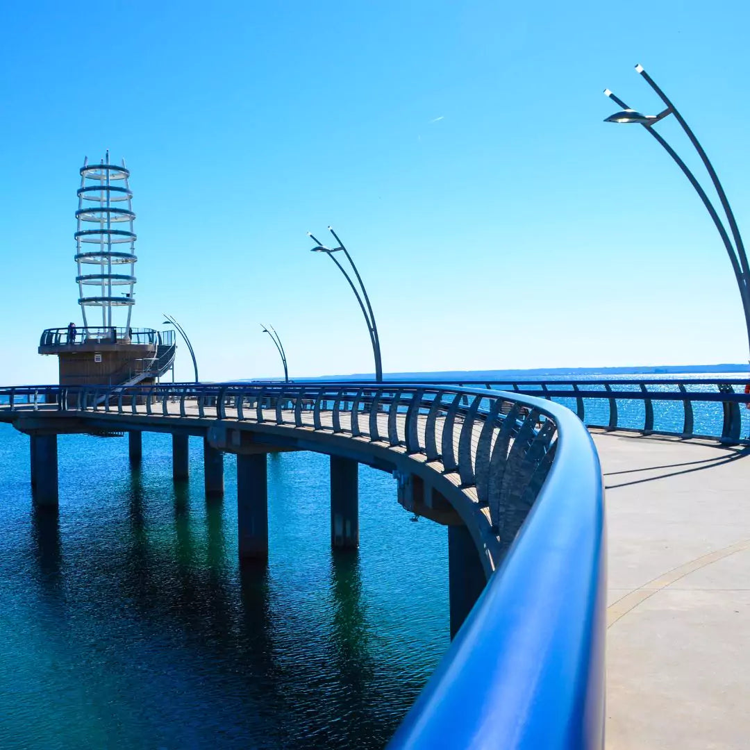 Curved boardwalk with modern streetlights extending over clear blue water beneath a bright sky.