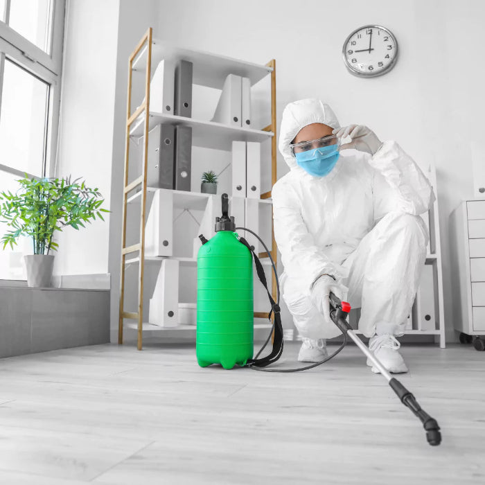 Wearing protective gear, a person kneels on the floor with Power HVAC Services Disinfectant Spray in Air Ducts in a room featuring shelves, a clock, and a potted plant.