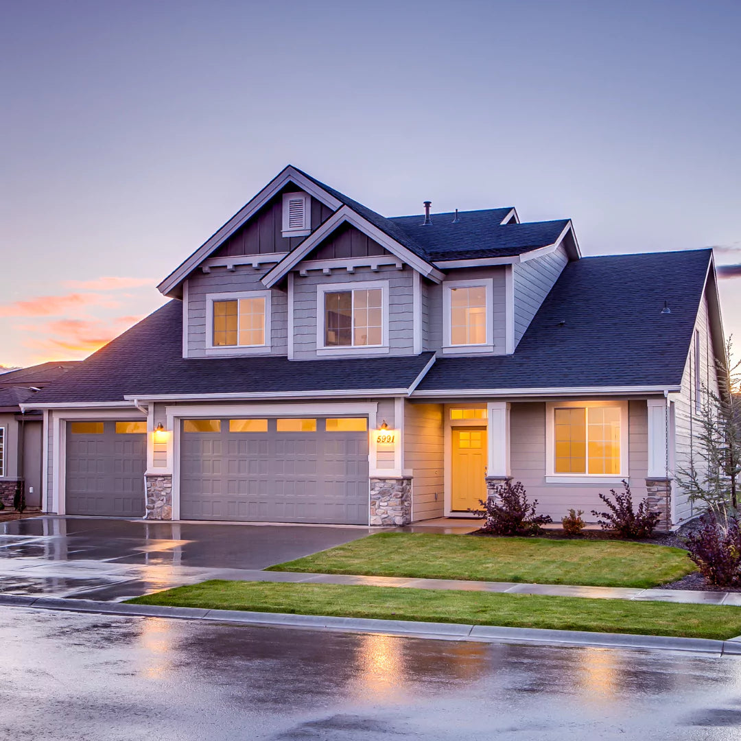 A two-story suburban house with gray siding, stone accents, and a three-car garage glows in the twilight. As rain kisses the pavement, rely on Power HVAC Services Air Duct Cleaning of Houses, following NADCA ACR 2021 standards, to keep your home as pristine inside as it looks outside.