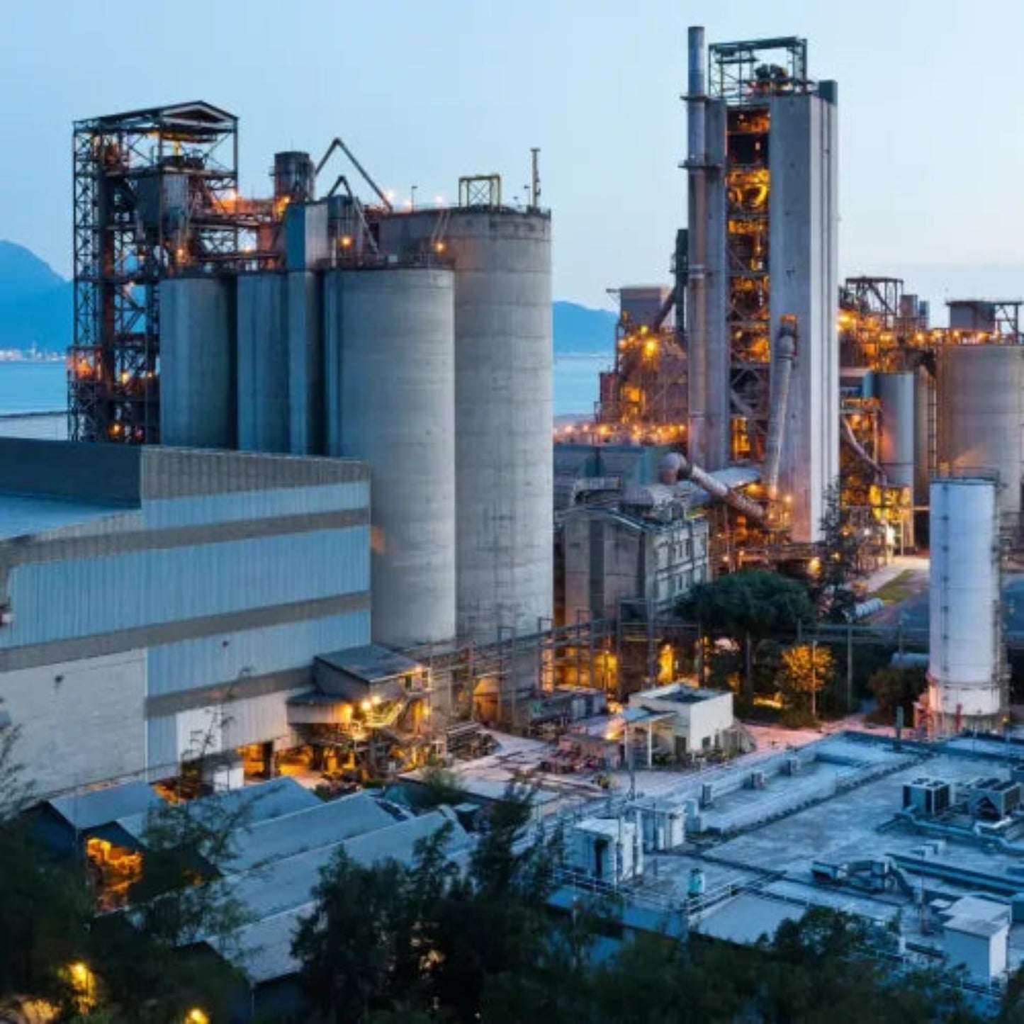 An industrial facility with large silos and metal structures is illuminated against a backdrop of mountains and a dimming sky. In the bustling GTA area, Power HVAC Services Industrial Air Duct Cleaning uses robot-assisted technology to ensure precise maintenance of this grand structure.